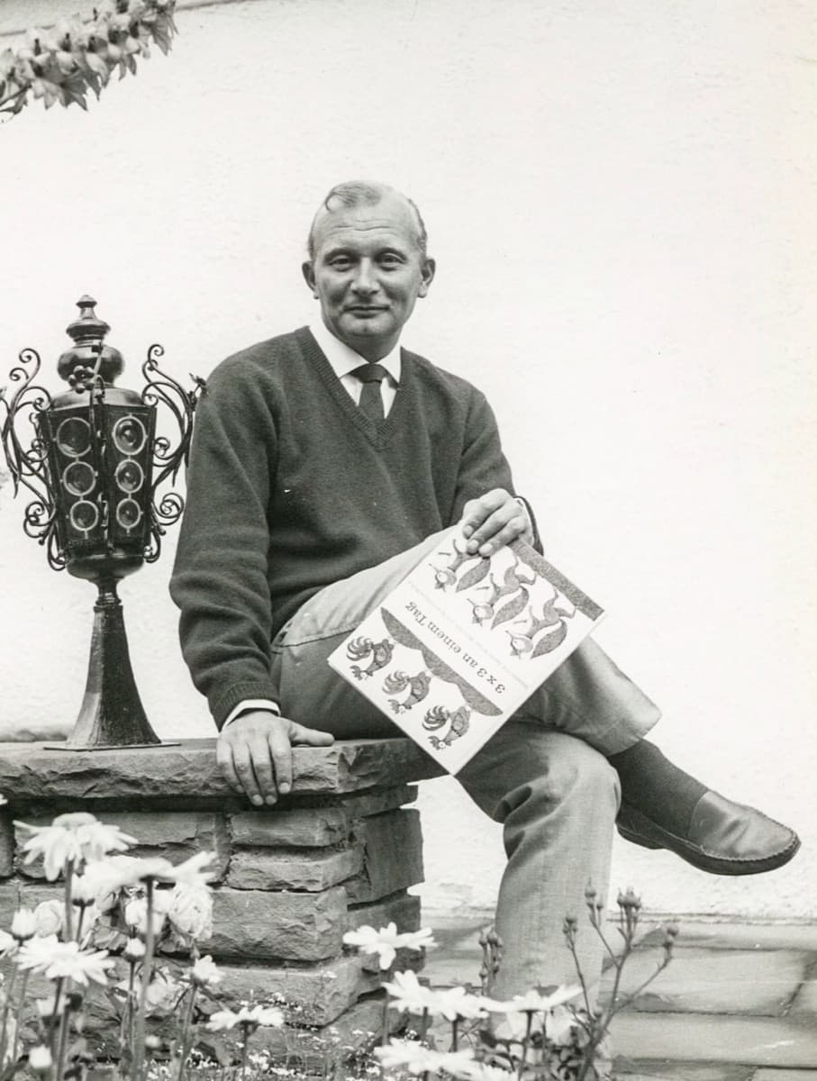 James Krüss, 1963, sitzt auf einer Steinmauer und hält ein Bilderbuch in den Händen. Schwarz-weiß-Aufnahme aus seinen Münchner Jahren.