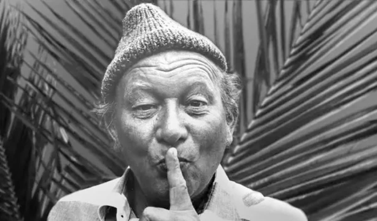James Krüss als älterer Mann, mit Wollmütze und dem Finger vor den Lippen, vor einem Palmen-Hintergrund. Schwarz-weiß-Porträt aus seinen Gran-Canaria-Jahren.