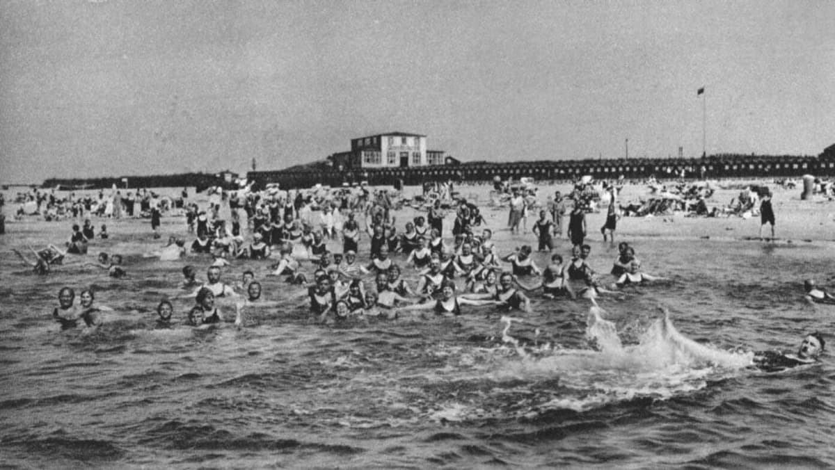 Historische Aufnahme von Badegästen am Helgoländer Strand. Frauen in langen Badekleidern, Männer in gestreiften Anzügen, Bademaschinen am Wasserrand.
