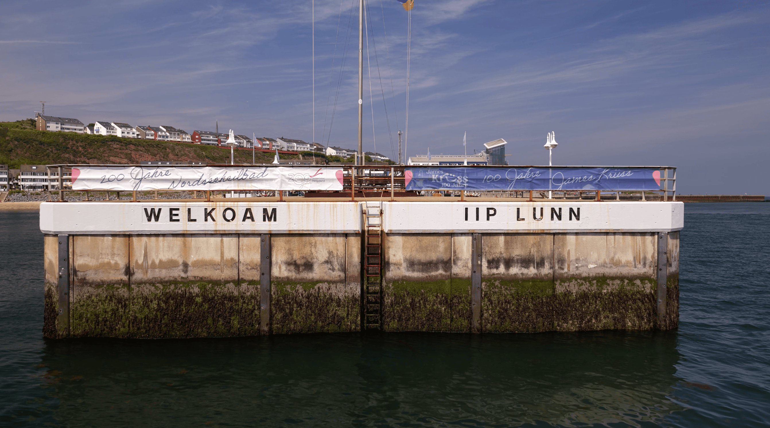 Stirnseite der Helgoländer Landungsbrücke vom Wasser aus. In weißen Lettern auf dem Beton: links 'WELKOAM', rechts 'IIP LUNN'. Auf der Brüstung darüber ein Banner '100 Jahre James Krüss'.